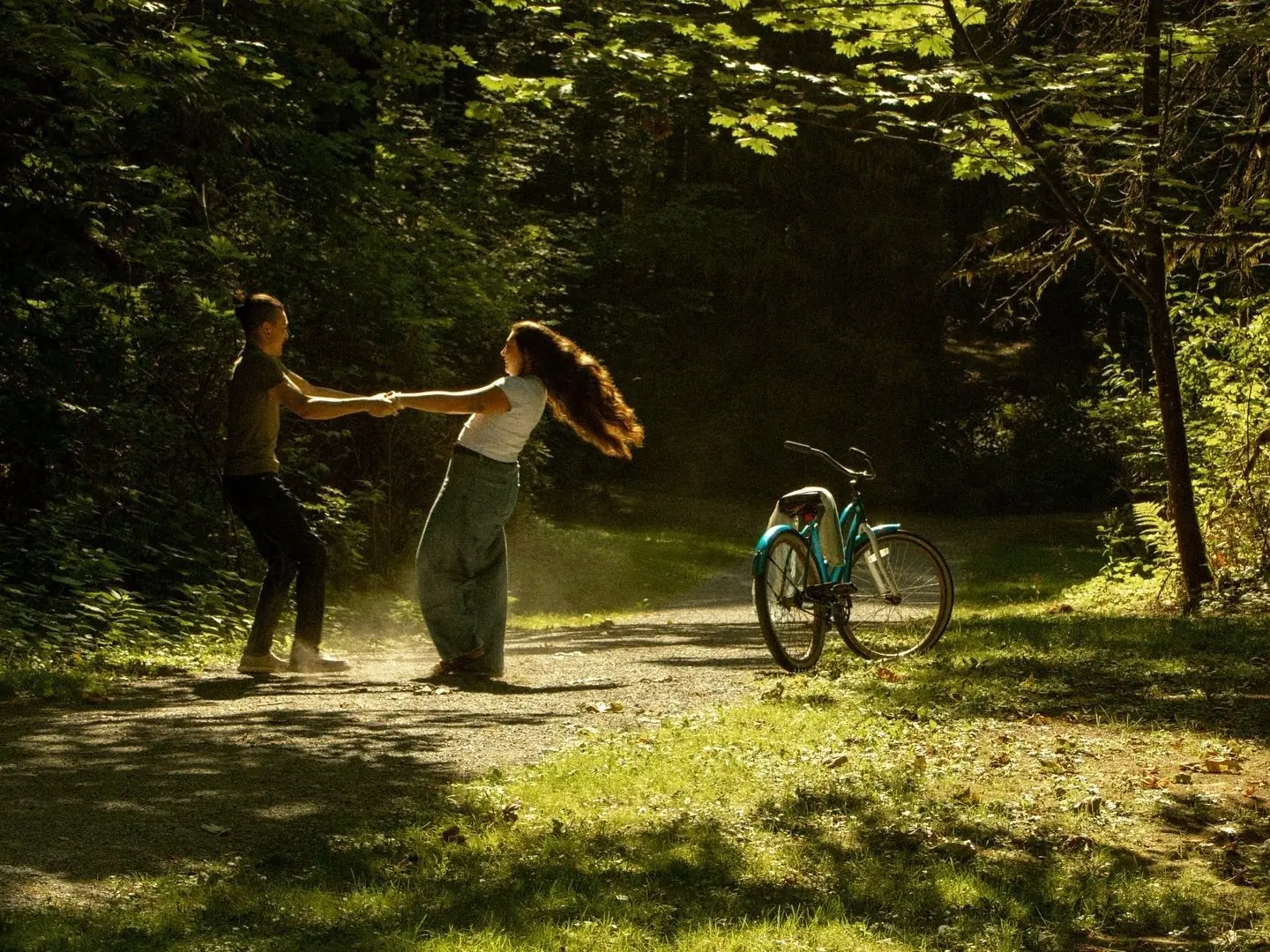 A couple dancing on a sunlit forest path beside a bicycle